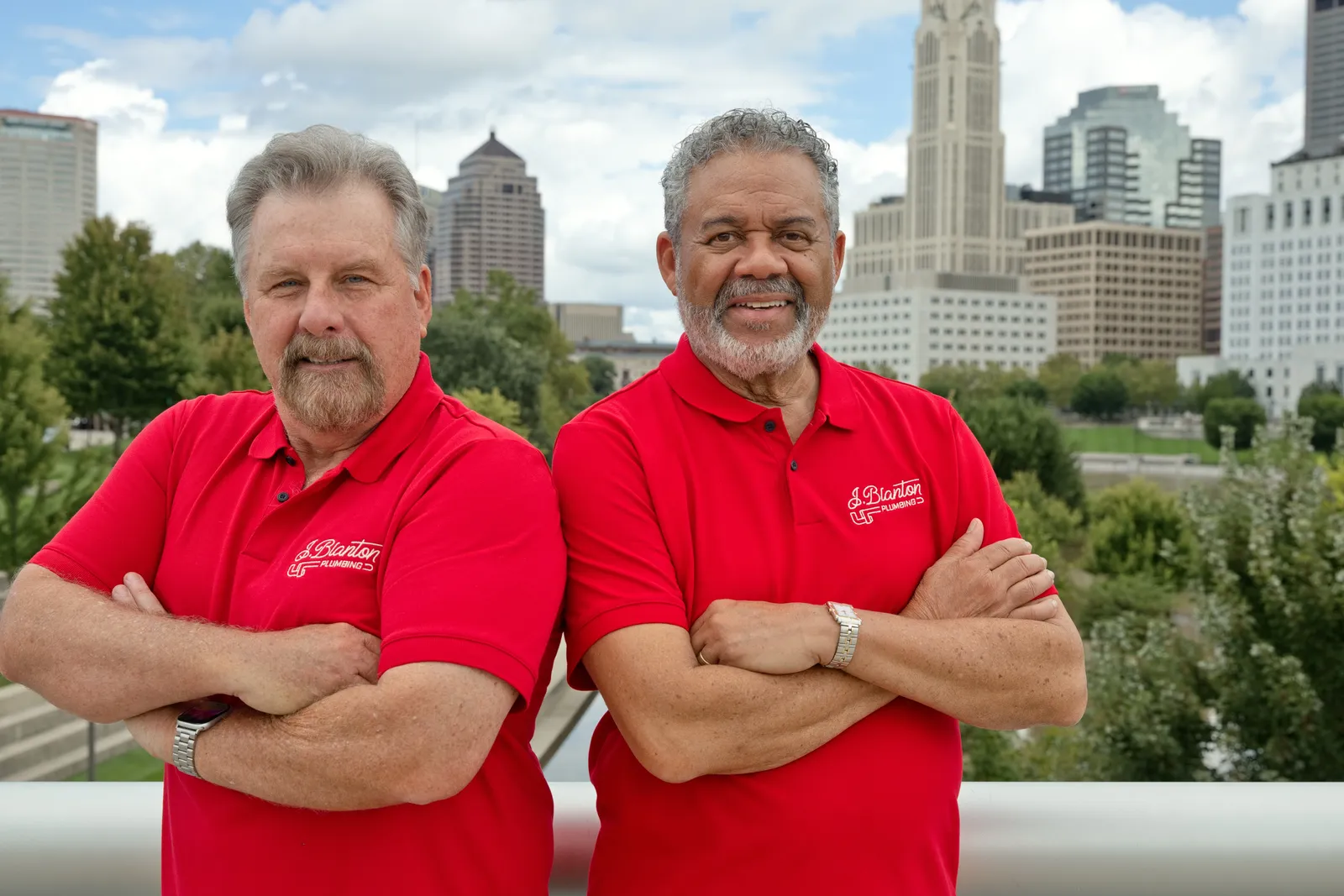 Two J. Blanton Plumbing team members in red company polos, arms crossed, in front of the downtown Columbus skyline