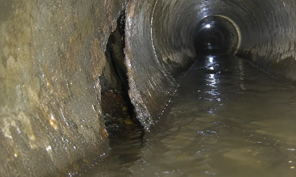 In-pipe camera view of a corroded sewer pipe wall with a visible crack, water flowing through the damaged section